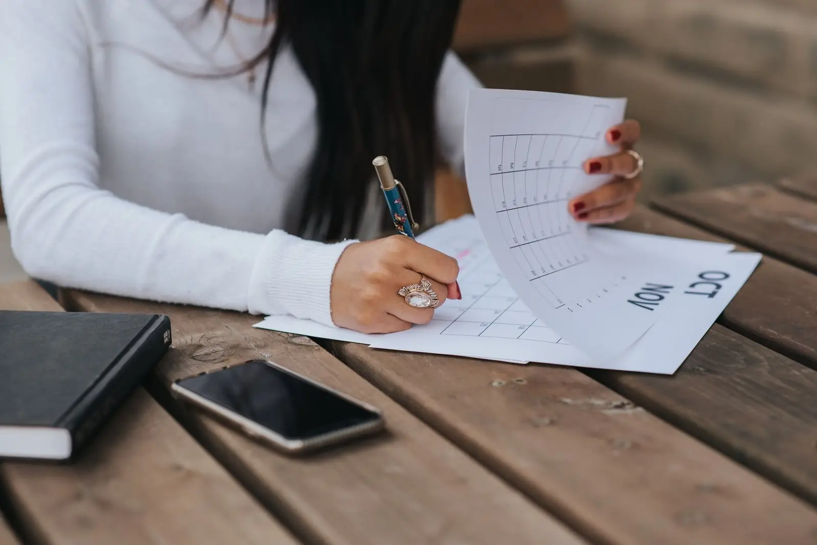 A person signing a printed form at a wooden table, with a notebook and phone beside the papers